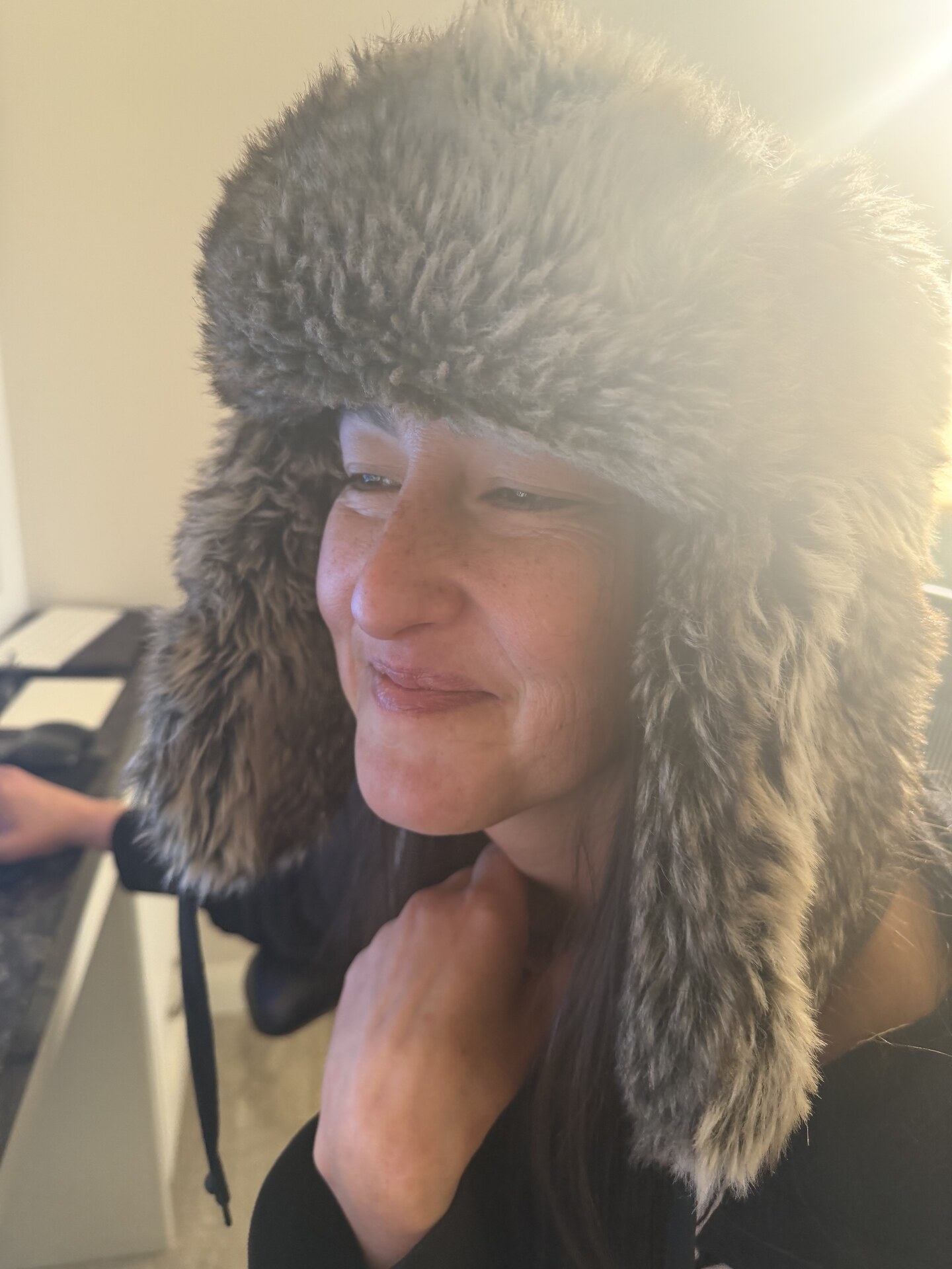 Jen in a fur-lined trapper hat, sunlight flaring behind her, looking off to one side.