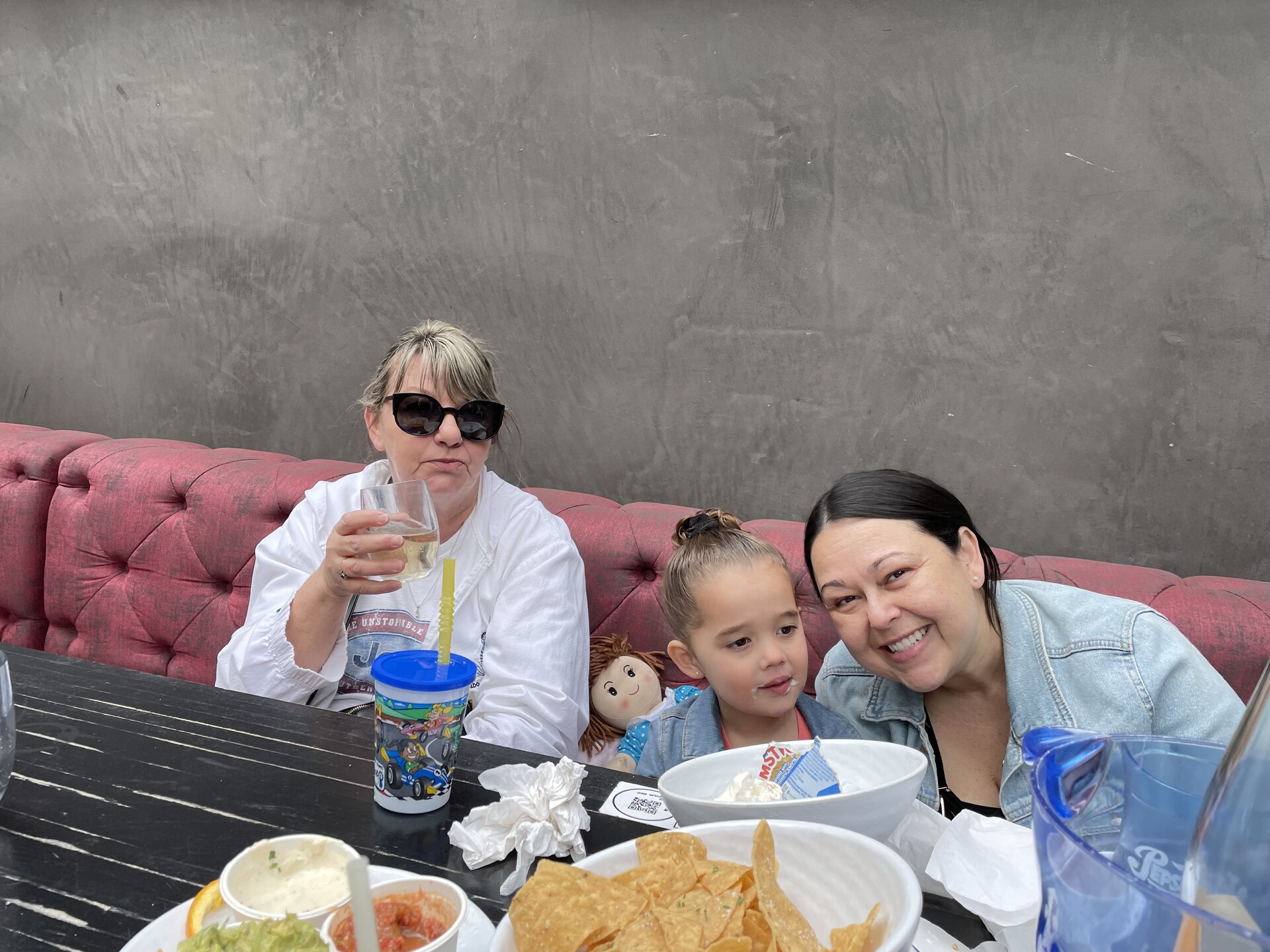 Two women and a young girl crowded into a pink restaurant booth behind bowls of chips and salsa, one of them raising a glass.