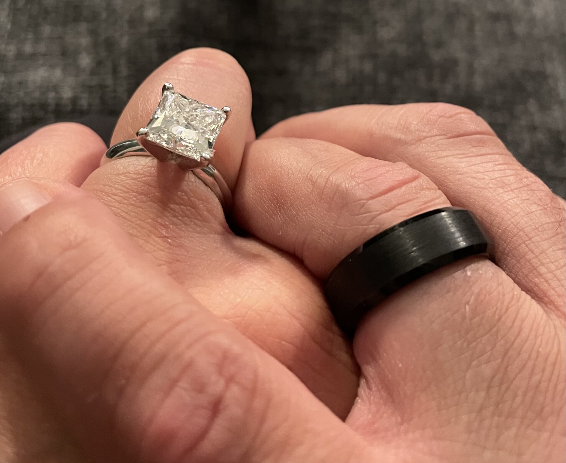 Two hands resting together — a princess-cut diamond on hers, a brushed black band on his.
